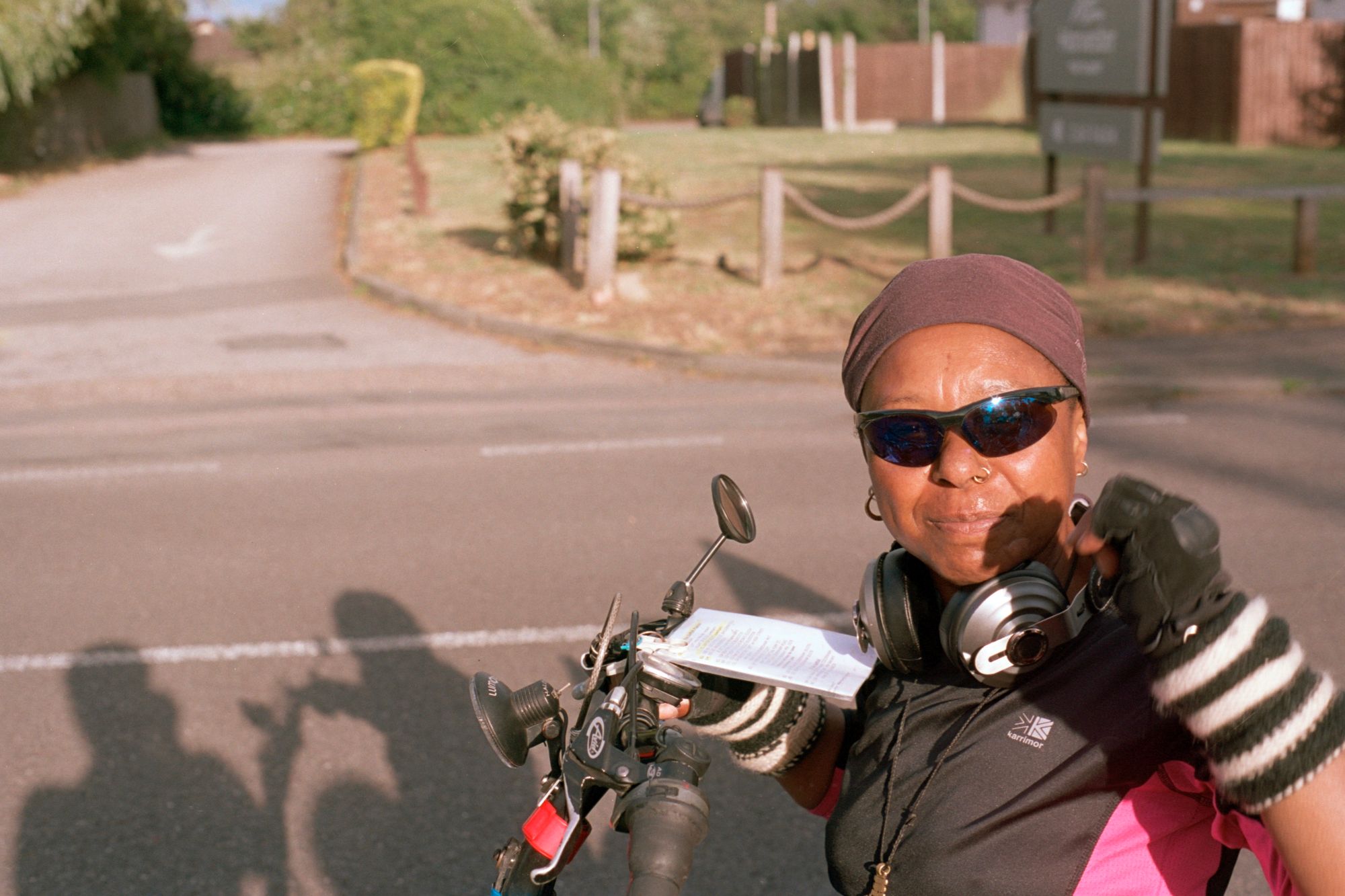 Denise, a Black woman wearing gloves, wrist protectors, sunglasses, nose ring and earrings, a Karrimor jersey, and a cross, with a pair of headphones around her neck, sits astride her recumbent bicycle with a route sheet attached to the handlebars.