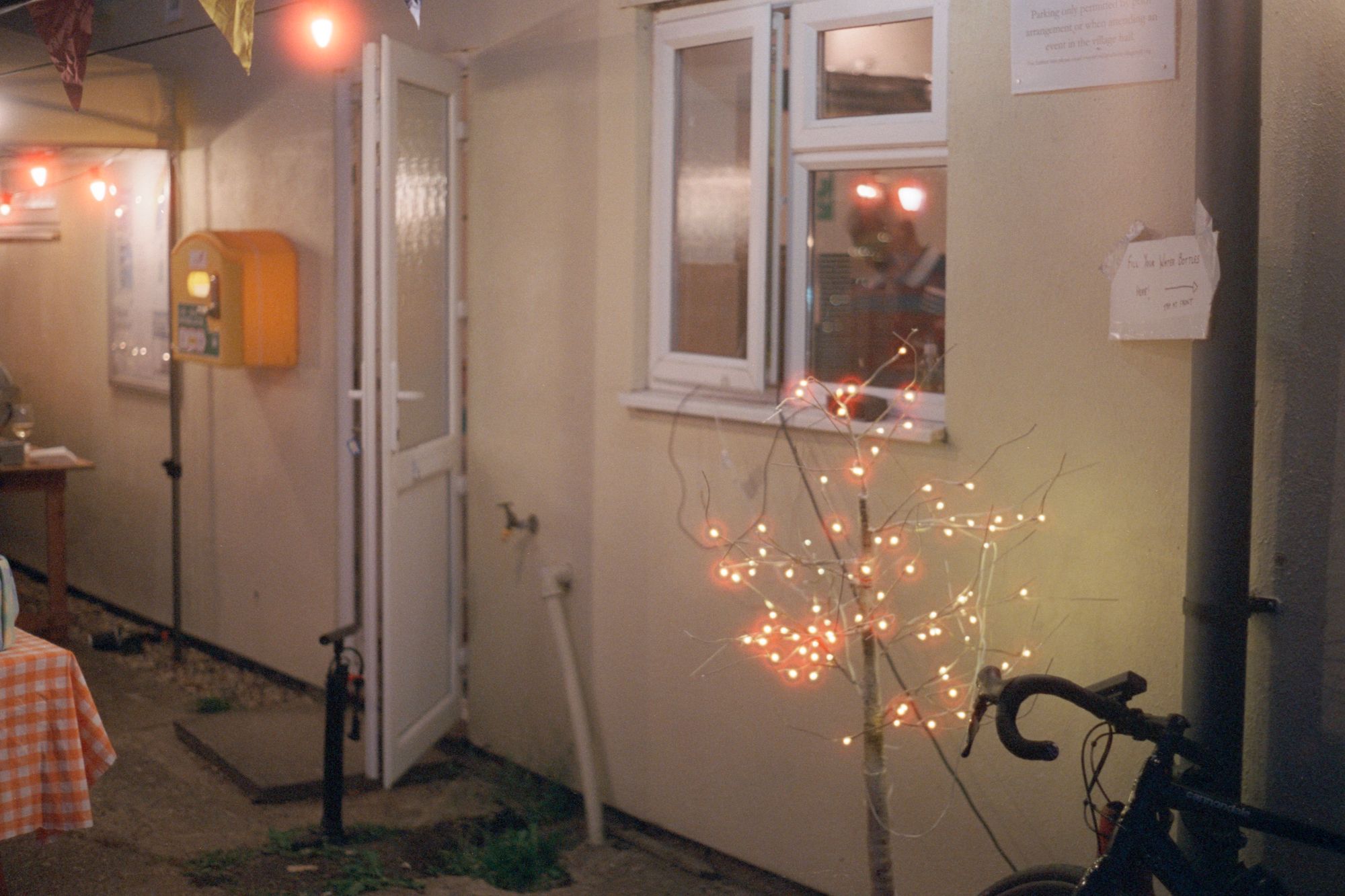 The wall of a community centre with one of those LED light trees in the foreground. In the foreground is the marquee festooned with bunting and string-candle lights, and the corner of a table with a gingham tablecloth. There's an automatic external defibrillator on the wall. A drop-bar bicycle stands against a drainpipe. A piece of paper taped to the drainpipe with an arrow pointing right reads: "FILL YOUR WATER BOTTLES HERE! TAP AT FRONT."