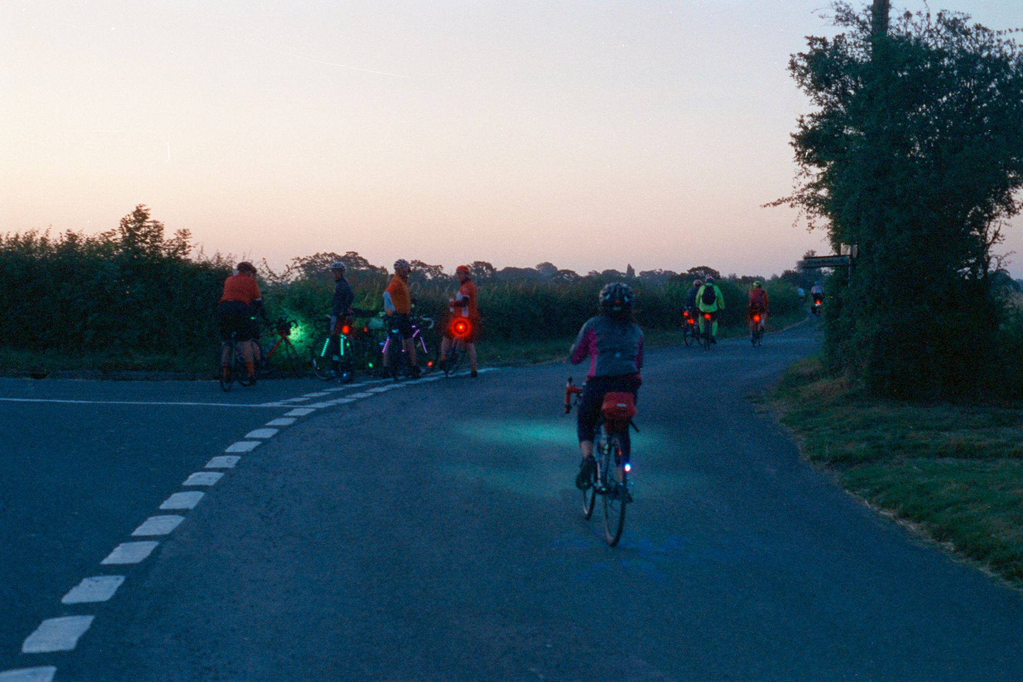 With another road joining from the right, cycling along a country road during a mauve-coloured dawn. Some people are stopped at the junction with green and purple strip LED lights on their bikes.