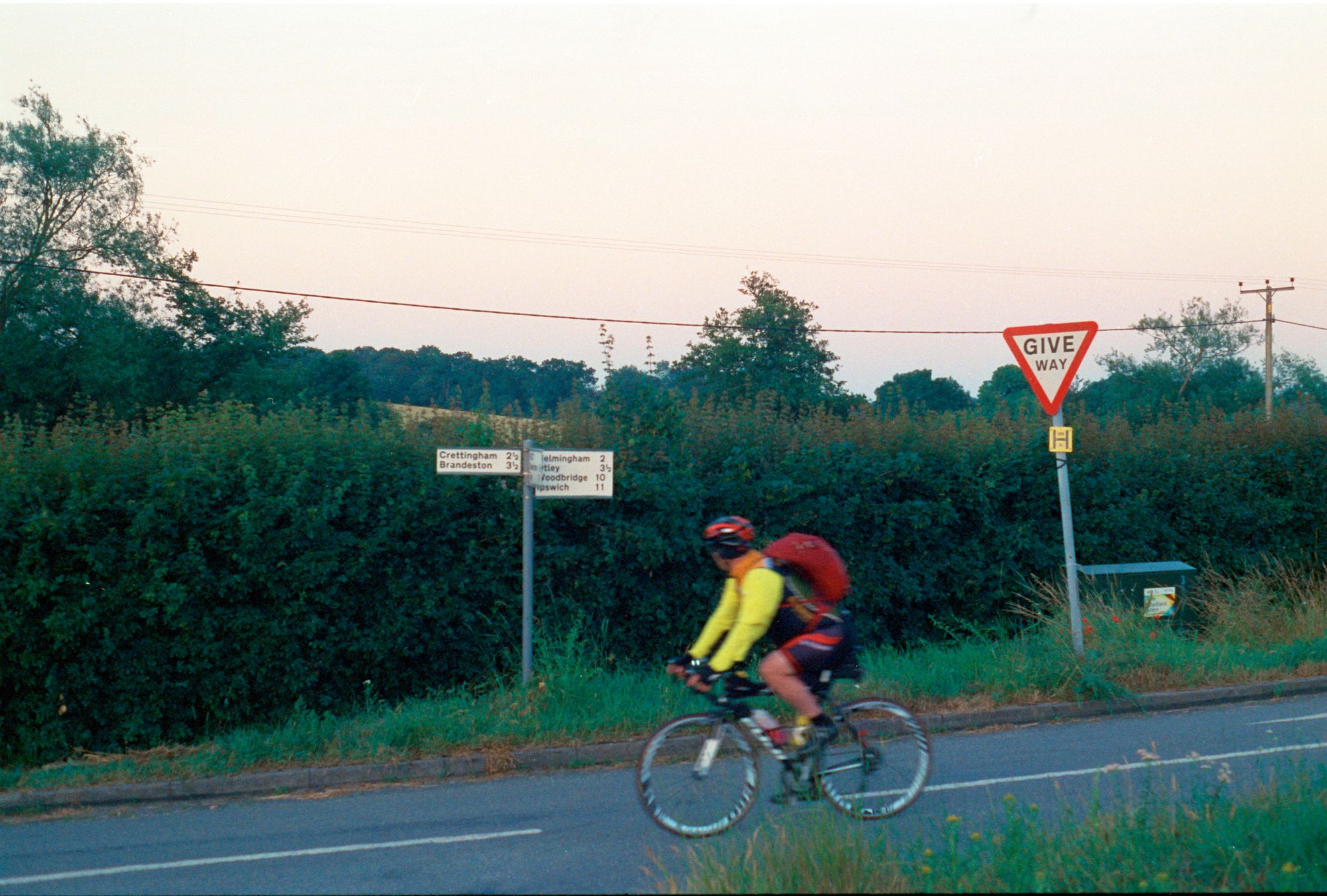 A cyclist in a fluorescent green jacket and with a red backpack on a road bike cycles right-to-left past a "GIVE WAY" sign and a signpost reading "Crettingham 2½ miles left, Brandeston 3 ½ miles left, Helmingham 2 miles right, Otley 3½ miles right, Woodbridge 10 miles right, Ipswich 11 miles right."