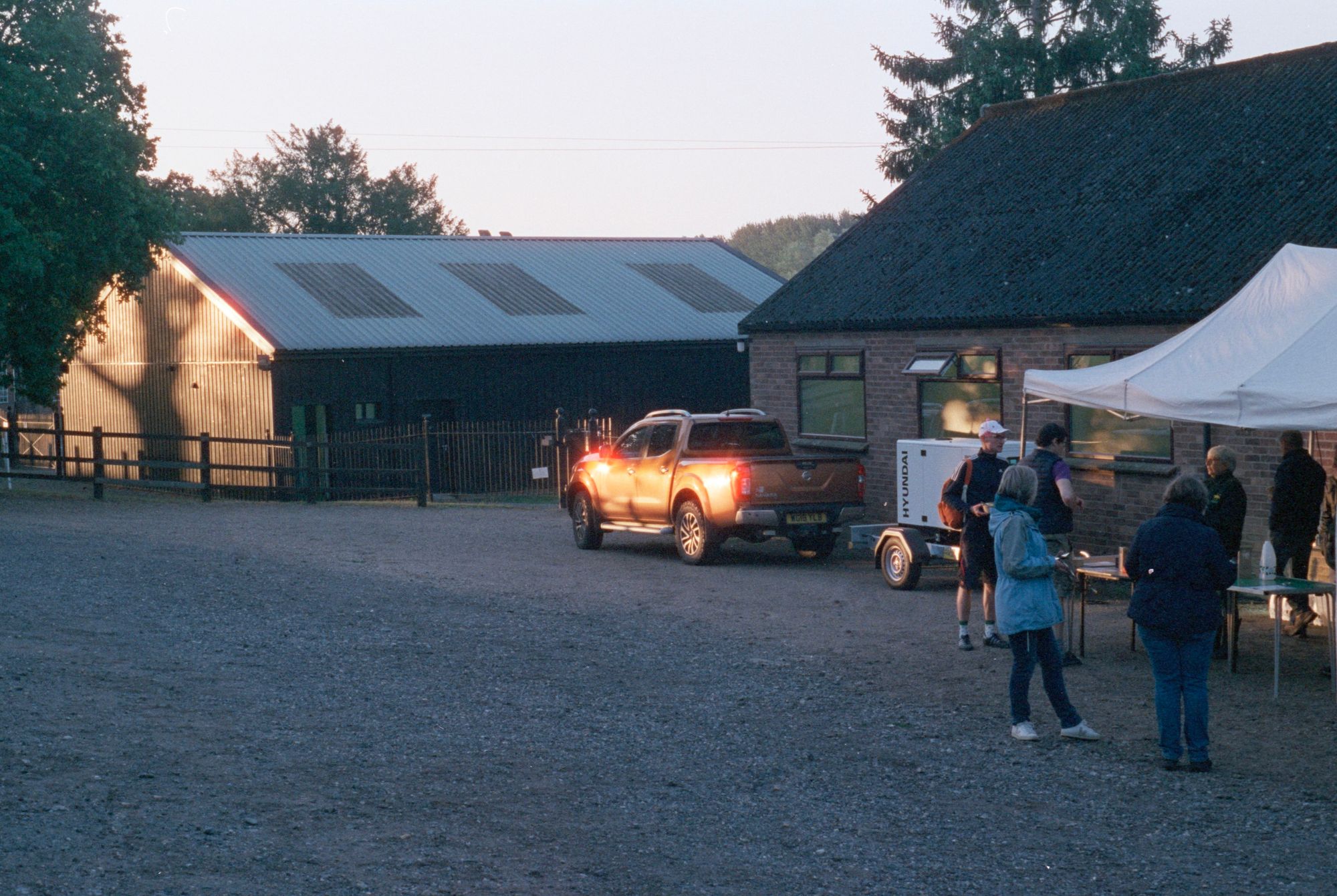 People line up and wait by a gazebo with a table. Sunlight shines on an orange pickup truck which glows in response, and a barn which has the shadow of a tree cast on its corrugated walls.