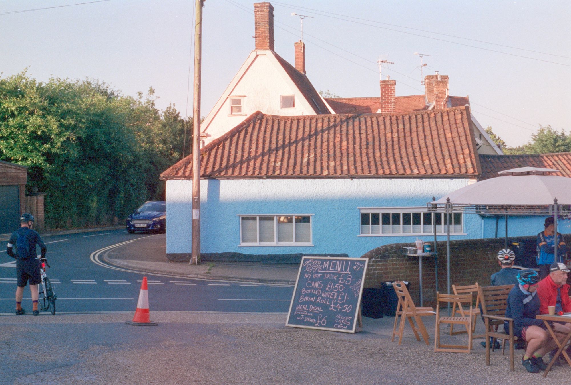 A car park in a village with wooden chairs and a blue building with the sun shining on it. Cyclists sit on the chairs. A chalkboard reads: "Menu: Crisps £1, soup £3+roll, cakes £2.50, any hot drink £3, Soup + Roll £3, Cans £1.50, Bottled water £1, Bacon roll £4.50, meal deal: Bacon roll + hot drink £6, accept cards + cash."