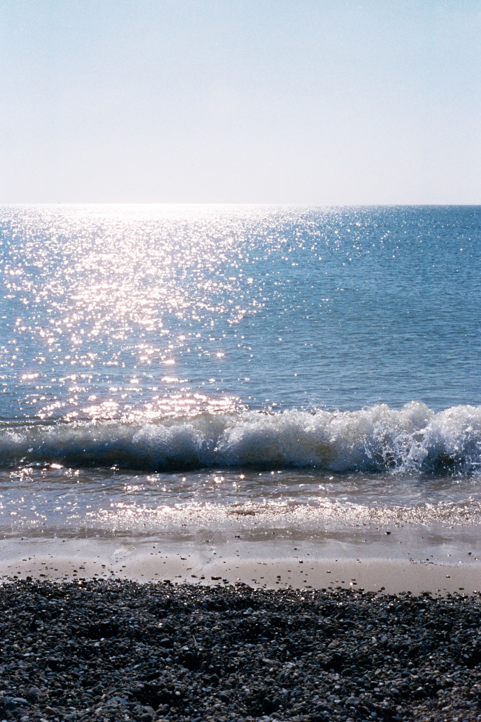 A view out to sea from a shingle beach, with the water and the gentle waves sparkling in the fresh morning blue.