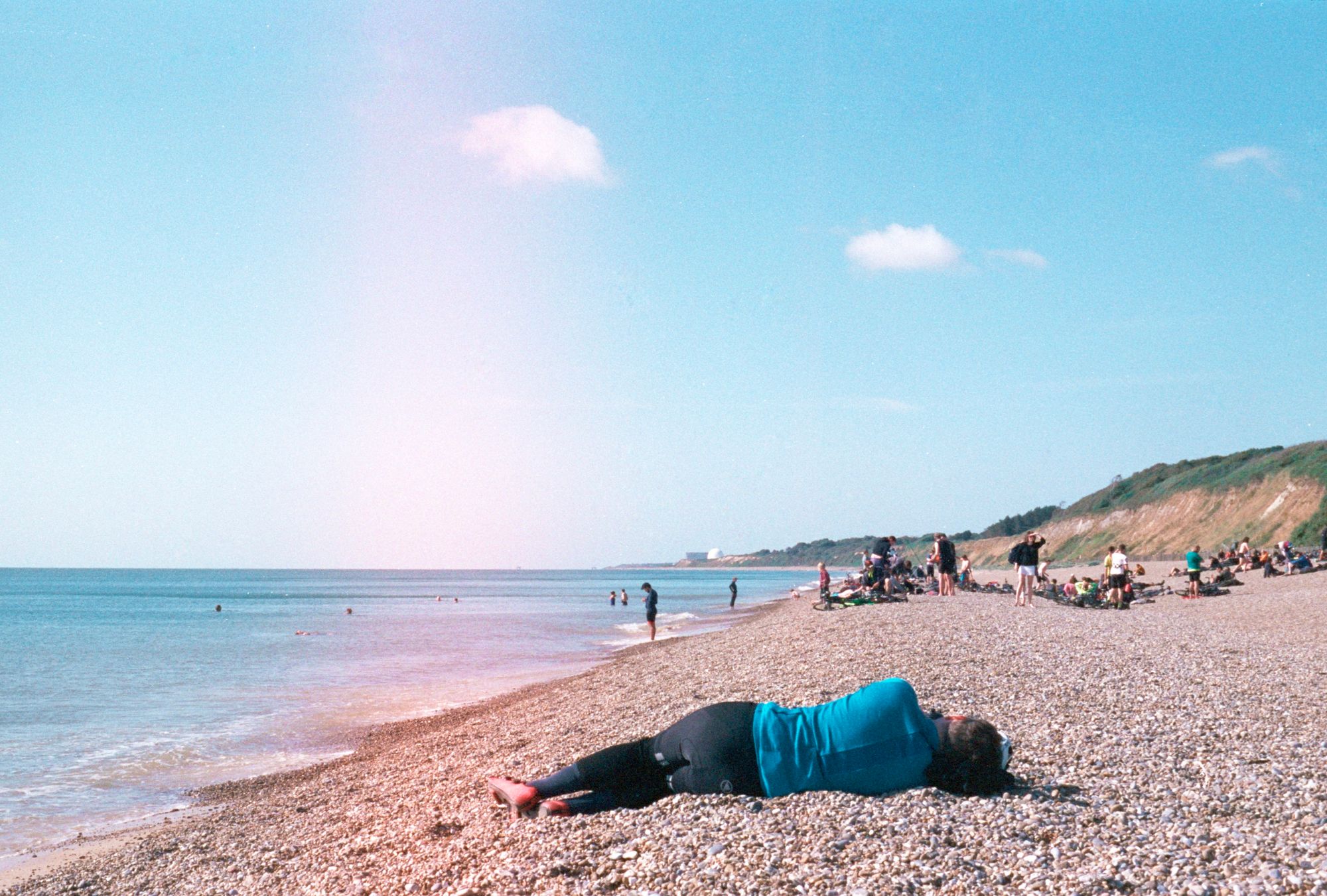 A cyclist lies on a shingle beach with people milling about near the sea and some swimming in the sea itself on a sunny day. In the distance, the white dome of Sizewell B power station sticks out on the sloped coastline like a sore thumb.