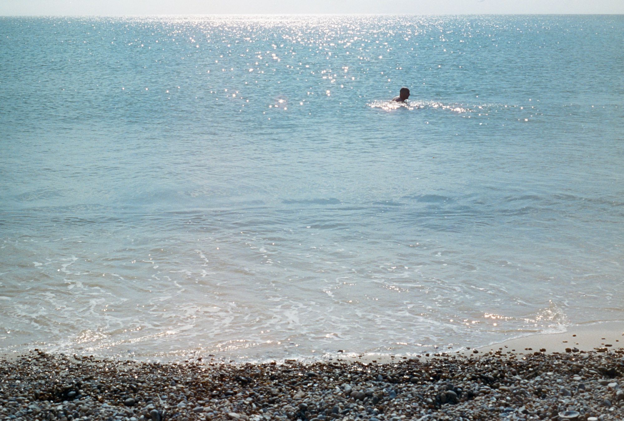 View out to the sparkling North Sea from the shingle Dunwich beach. A man with cropped hair in silhouette swims left to right around 10 metres out to sea.