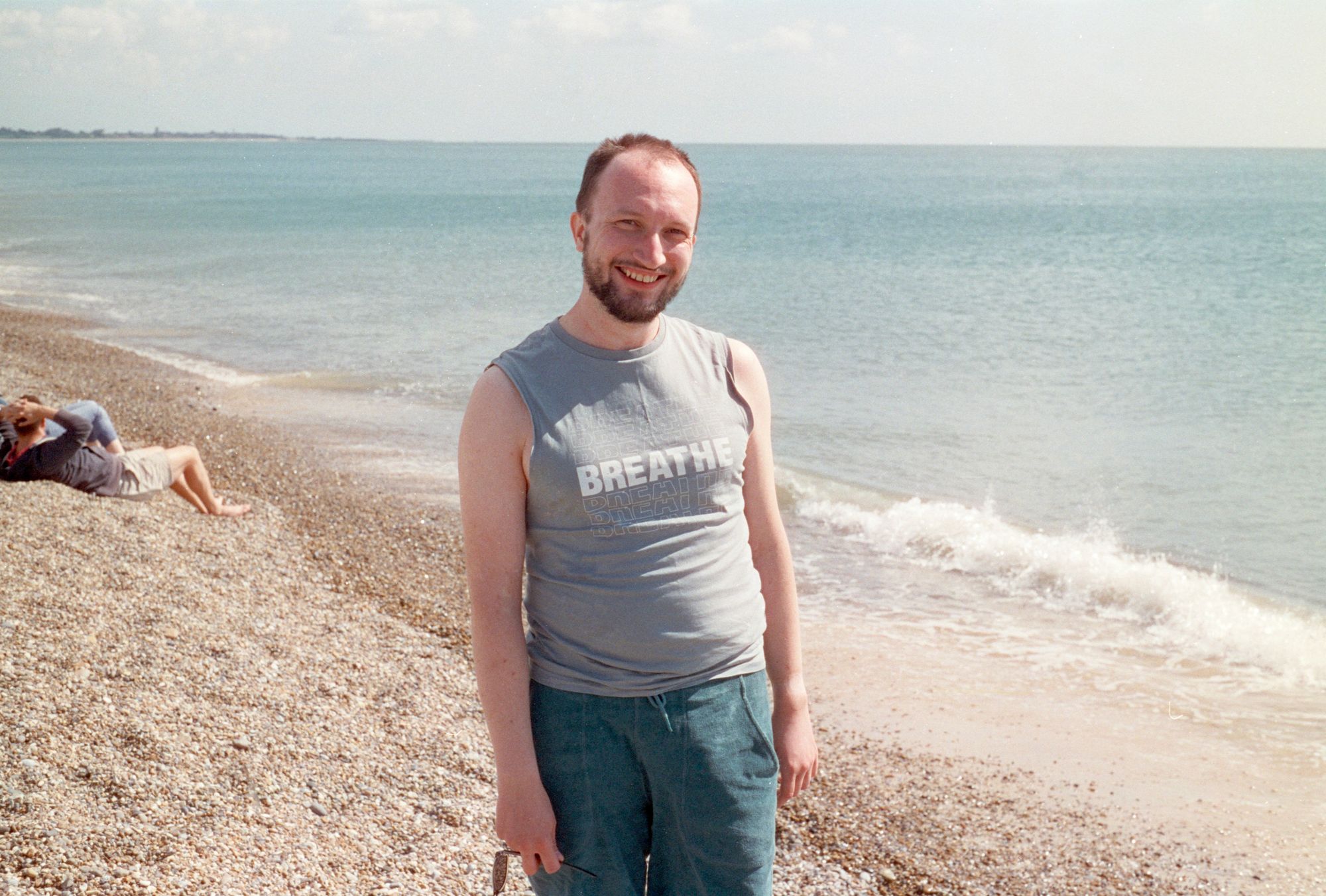 Jonathan, a white man with short hair and a scruffy beard, standing on the beach, facing camera, smiling. I'm wearing green towelled shorts and a teal-coloured vest with the word BREATHE on the front.