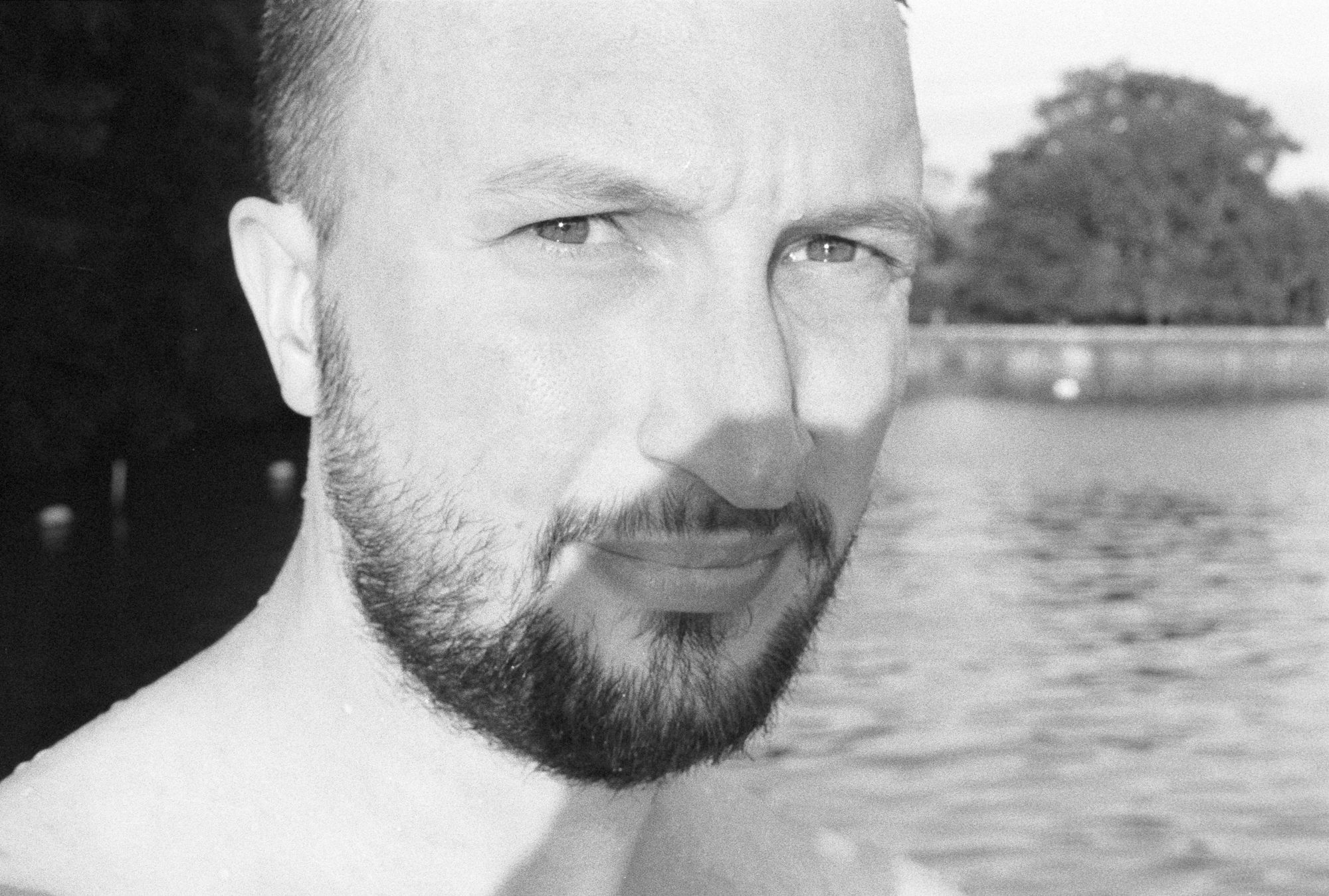 Head and shoulders shot of Jonathan on black-and-white, shirtless and with spots of water on his shoulders, staring into the sun (the camera casting a shadow over his beard) while standing in front of a swimming pond in a park.