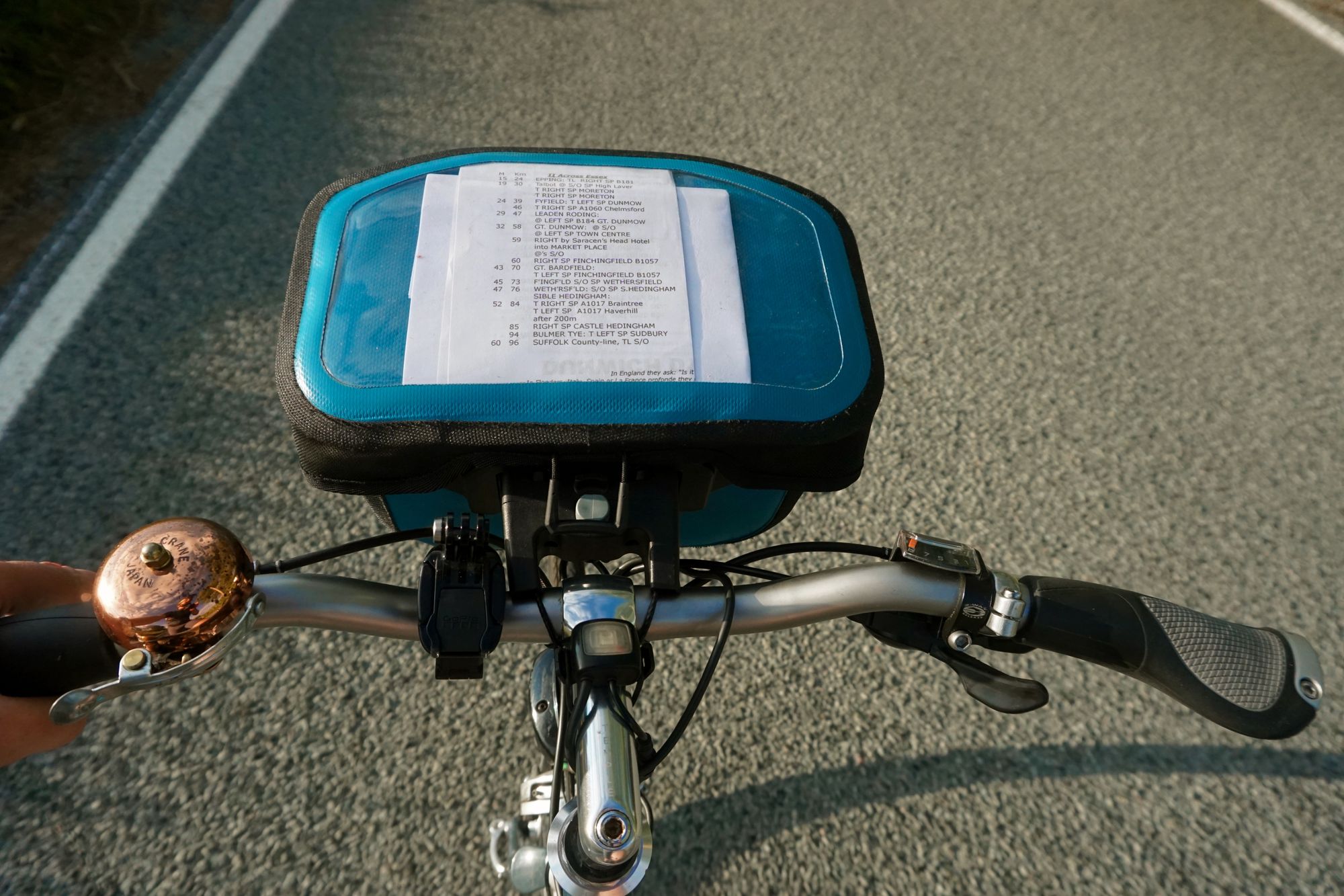 Shot from the saddle of a flat-handlebar bicycle's cockpit with a bronze Crane bell, rubber grips, and a blue handlebar bag. Inside the bar bag is a route sheet. It reads: II Across Essex 15 miles 24km EPPING: TL RIGHT SP B181 19 miles 30 km Talbot @ S/O SP High Laver T RIGHT SP MORETON T RIGHT SP MORETON 24 miles 39 km FYFIELD: T LEFT SP DUNMOW 46 km T RIGHT SP A1060 Chelmsford 29 miles 47 km LEADEN RODING: @ LEFT SP B184 GT. DUNMOW 32 miles 58 km GT. DUNMOW: @ S/O @ LEFT SP TOWN CENTRE 59 km RIGHT by Saracen's Head Hotel into MARKET PLACE @'s S/O 60 km RIGHT SP FINCHINGFIELD B1057 43 miles 70 km GT. BARDFIELD: T LEFT SP FINCHINGFIELD B1057 45 miles 73 km F'INGF'LD S/O SP WETHERSFIELD 47 miles 76 km WETH'RSF'LD: S/O SP S.HEDINGHAM SIBLE HEDINGHAM: 52 miles 84 km T RIGHT SP A1017 Braintree T LEFT SP A1017 Haverhill after 200m 85 km RIGHT SP CASTLE HEDINGHAM 94 km BULMER TYE: T LEFT SP. SUDBURY 60 miles 96 km SUFFOLK County-line, TL S/O