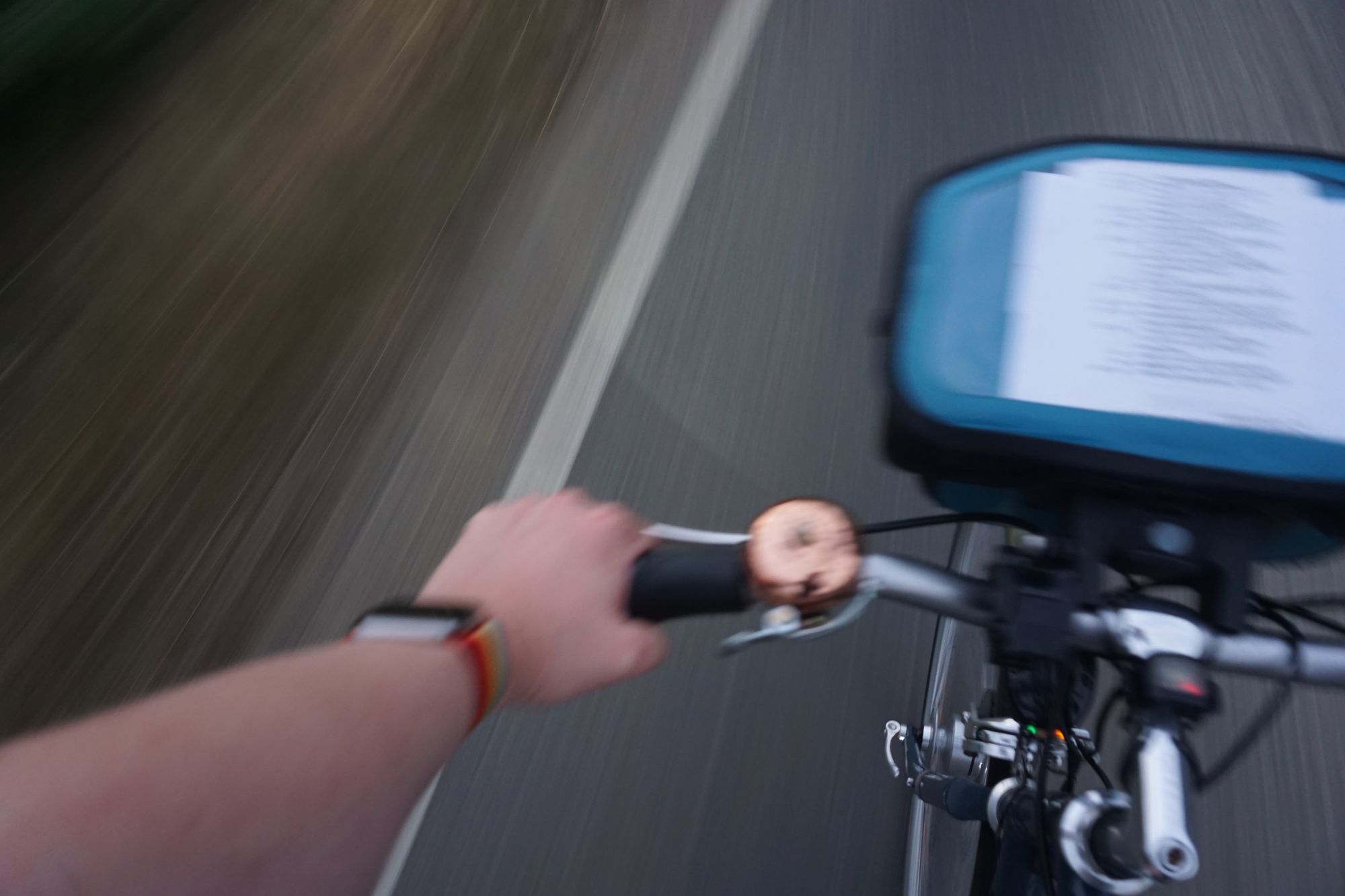 In motion: Point of view looking down on my left arm holding the left-hand grip on my bicycle's handlebar, with the route sheet visible on the bar bag and my Apple Watch with a rainbow strap. The bicycle's front light casts a pleasant curved cone of light ahead onto the road surface.