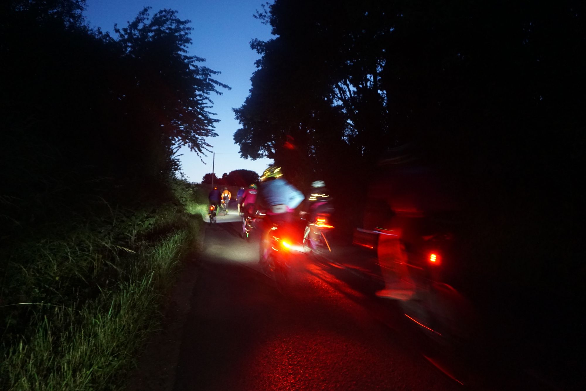 People cycling with glowing rear lights and front lights, and some people festooned in fairy lights, down an unlit country lane. In the distance, the pale blue of dawn rises beyond the road's edge.