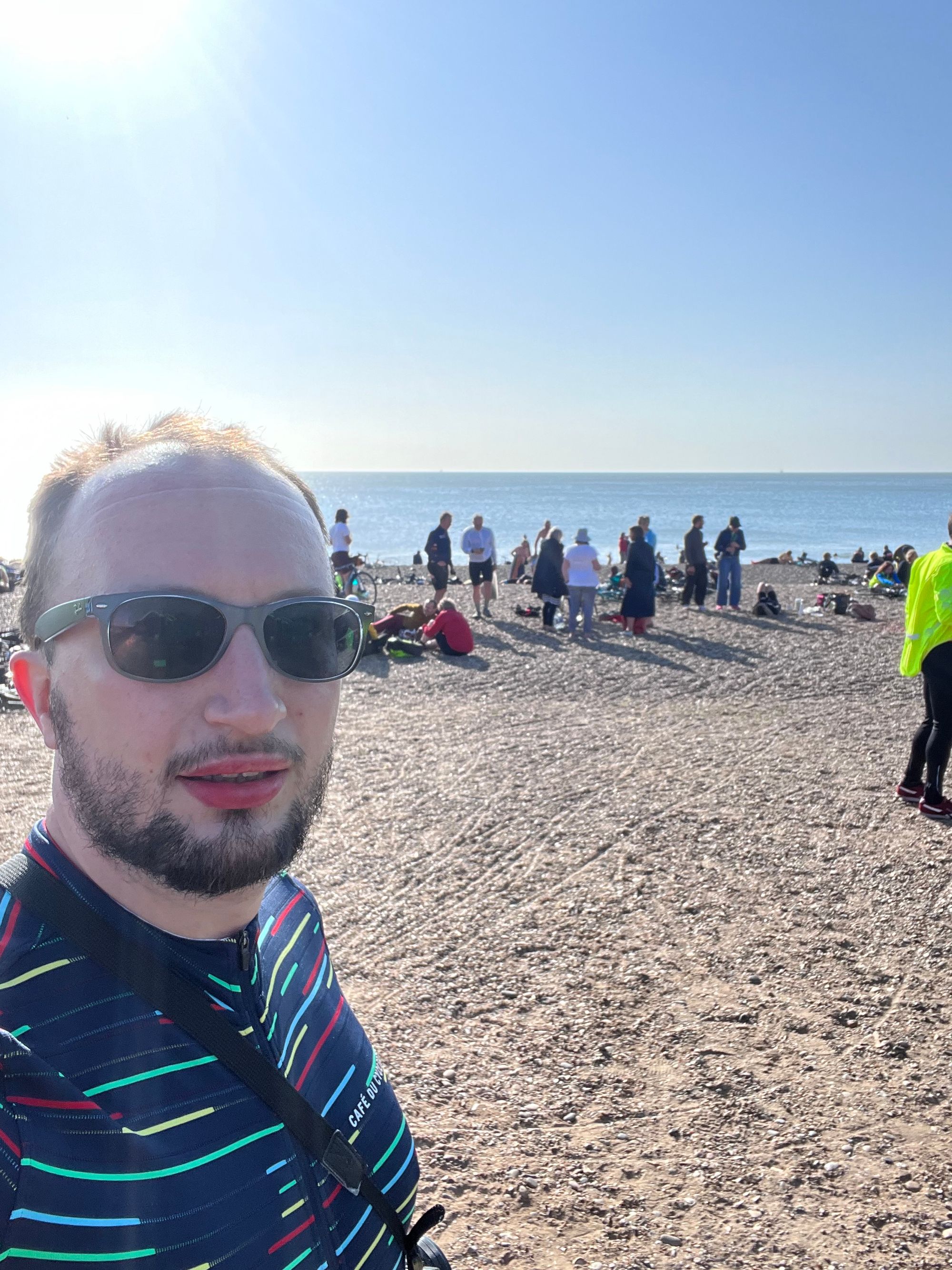 Selfie of Jonathan in sunglasses wearing a stripy cycling jersey waiting in a queue on the beach.