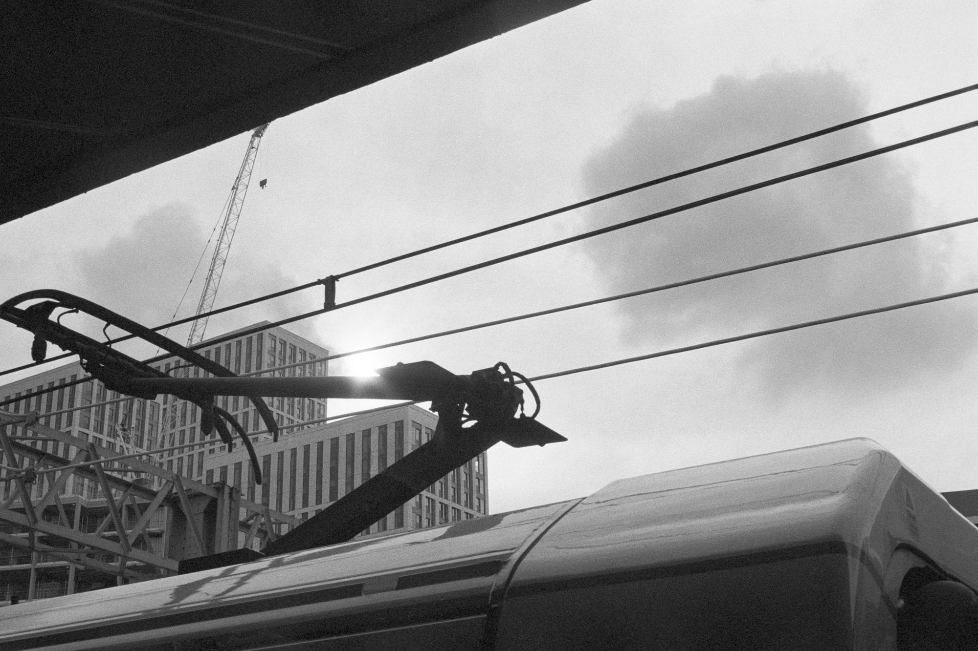 From a railway station platform, the gap between the platform canopy and the roof of an electric train. The pantograph is sprung into contact with the overhead electrical wires, through which the sun snaps through the clouds. In the distance, we see medium- and high-rise tower blocks of flats that look like cheaply constructed and architecturally non-descript cuboids, and an overhead gantry for the railway line.