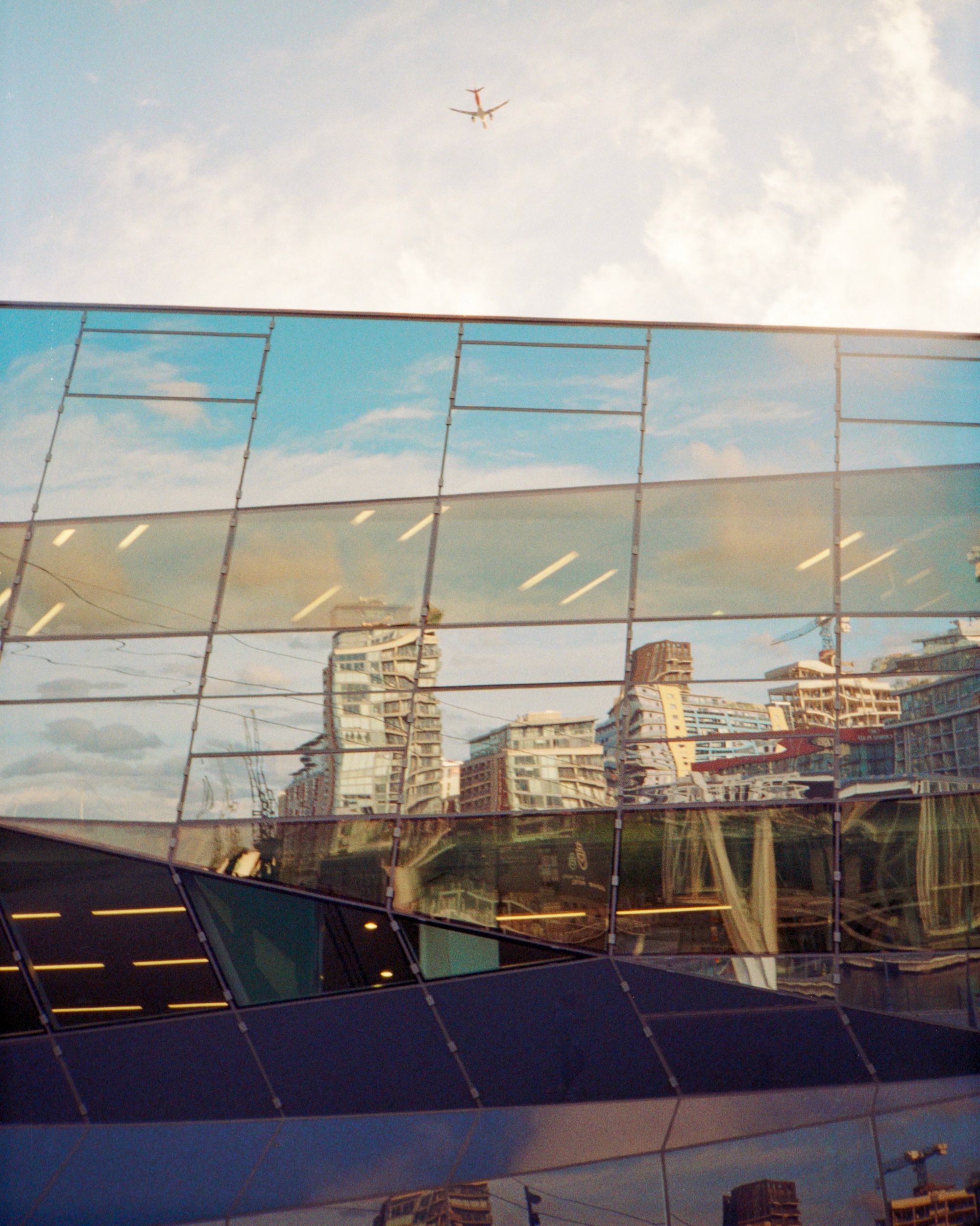 A skyline with a cablecar is reflected in a crystalline building with glass square panels that seem to crumple into triangular faces. Above, an aeroplane flies overhead on a blue, sunny day.