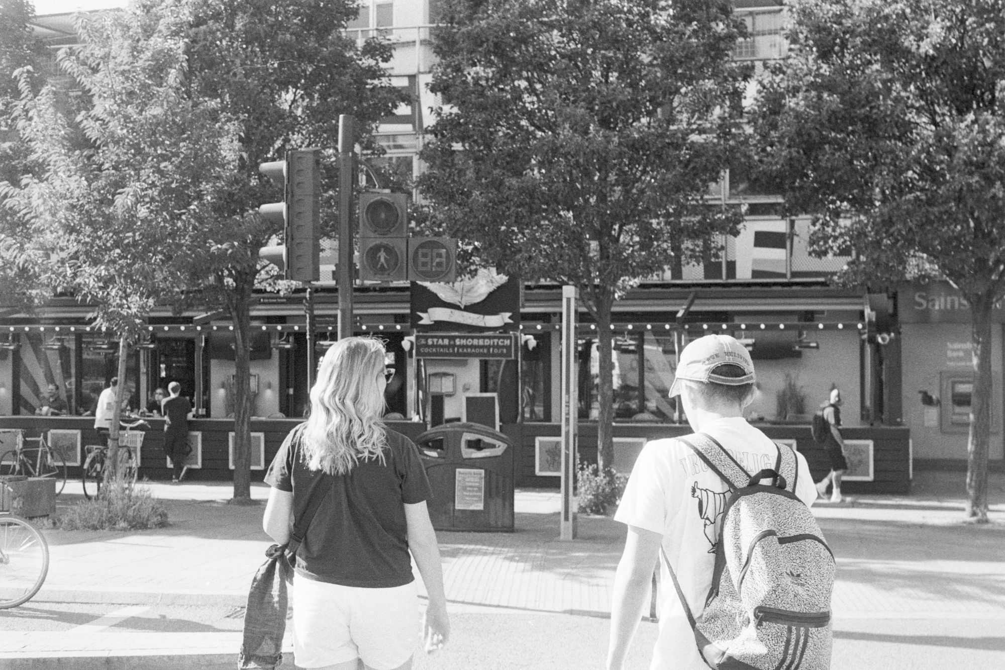Two people (a woman in a black t-shirt carrying a handbag and a man in a white t-shirt and cap) walk away from camera on a pedestrian crossing, towards a parade of shops with trees, in crisp black and white on a swelteringly sunny afternoon