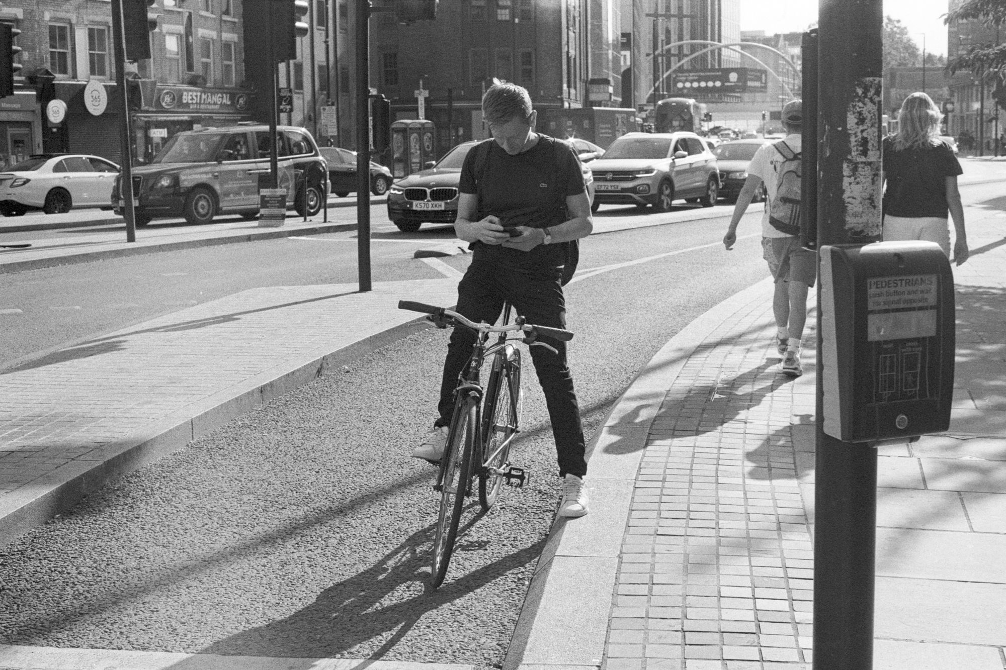 From a pedestrian crossing, a man in a black t-shirt on a bicycle sits waiting at the traffic lights, checking his phone, casting harsh diagonal shadows
