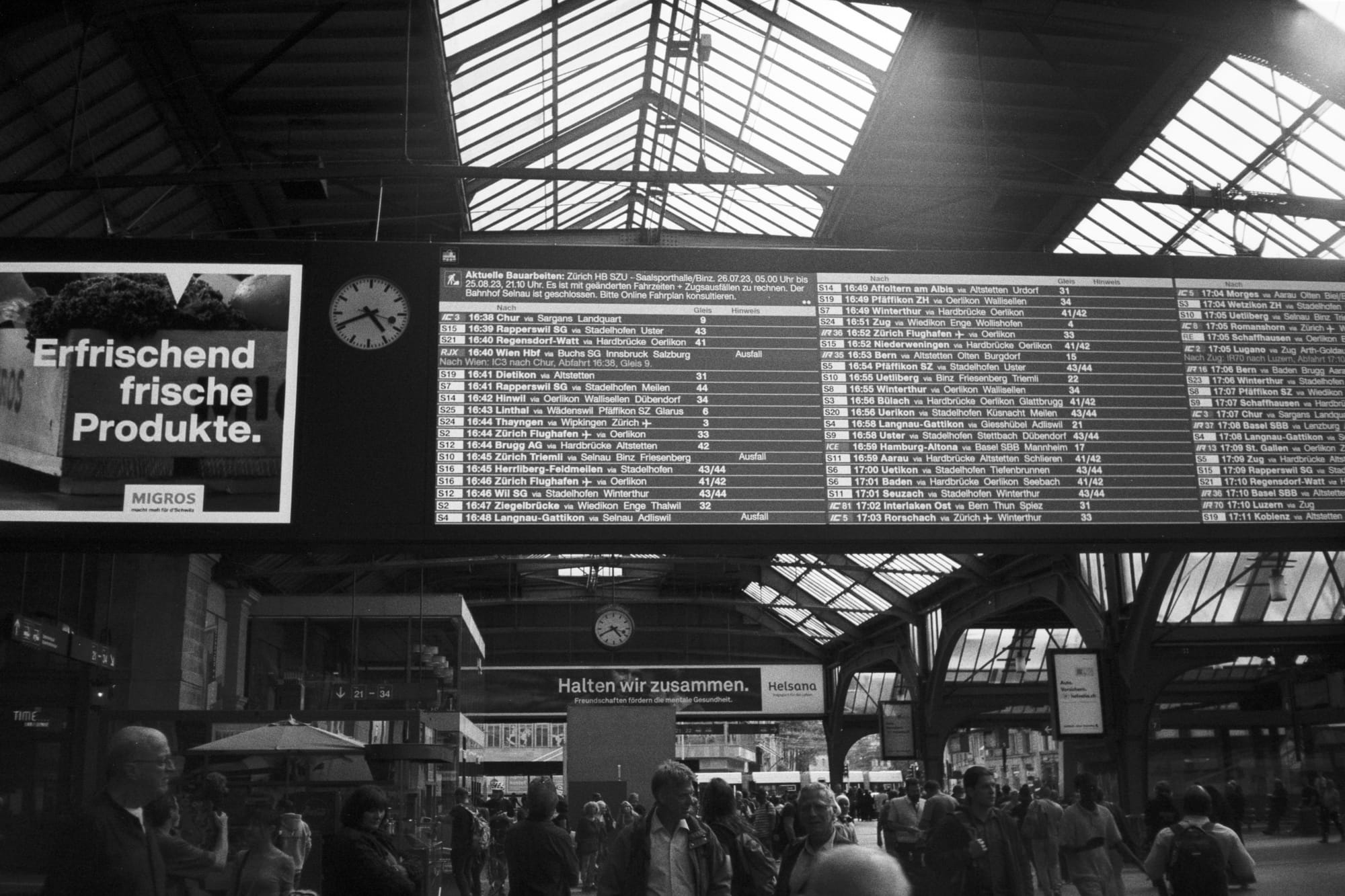 In black and white, a large railway station departures board for S-Bahn and ICE trains, in German. People bustle about the departures board below.