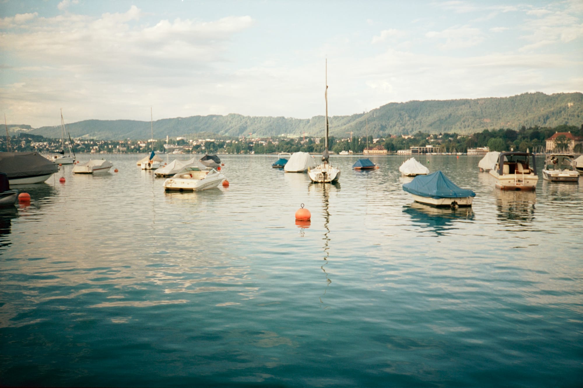 Lake Zürich, a clear, stunning blue. Small boats, yachts, and buoys rest on the surface. The sky is blue and fluffy. All is peaceful.