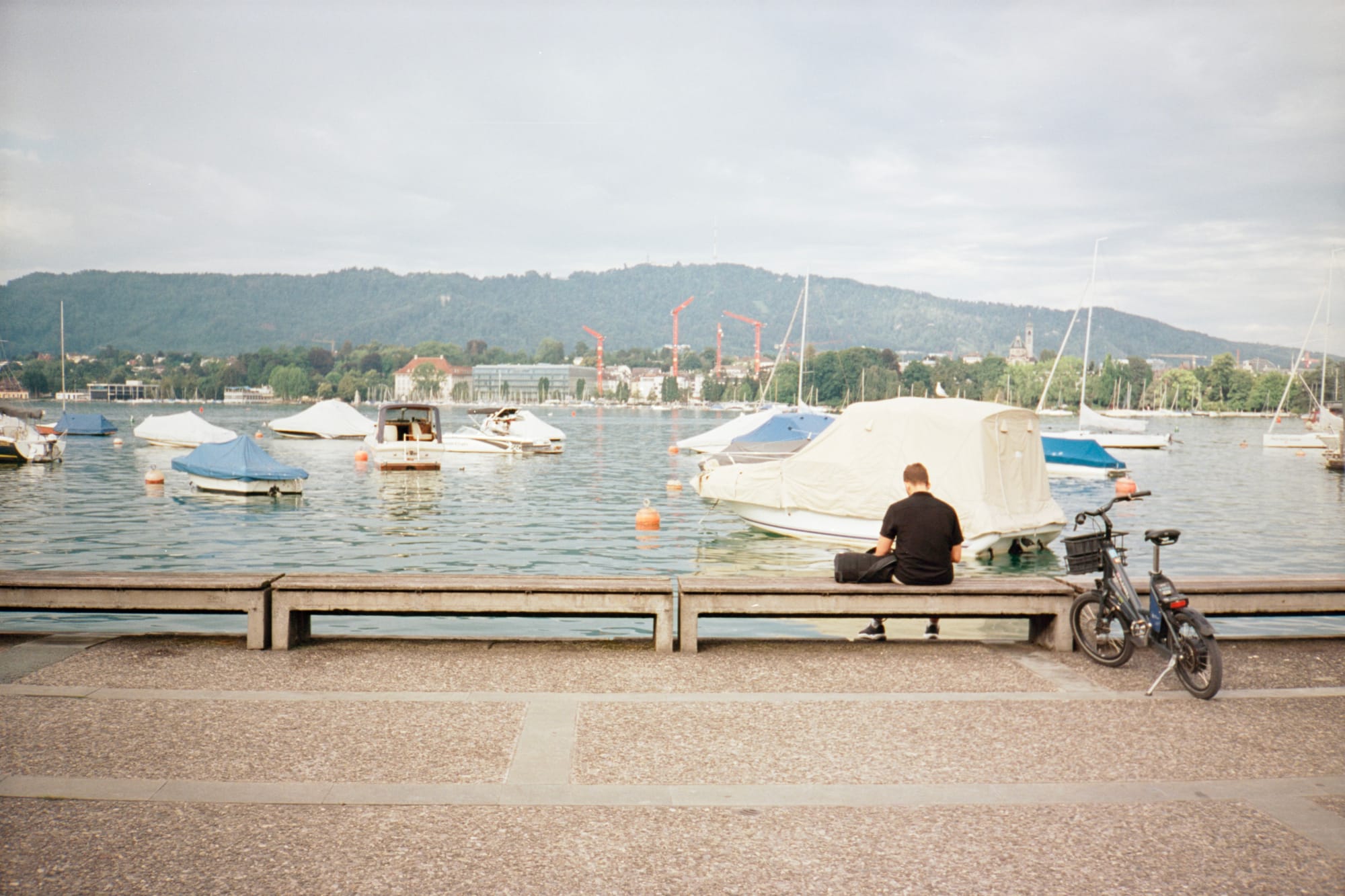 A man in a black t-shirt sits on a bench facing out onto Lake Zürich, reading his phone. His small electric bicycle is on a kickstand nearby. On the lake are tons of small boats and buoys.