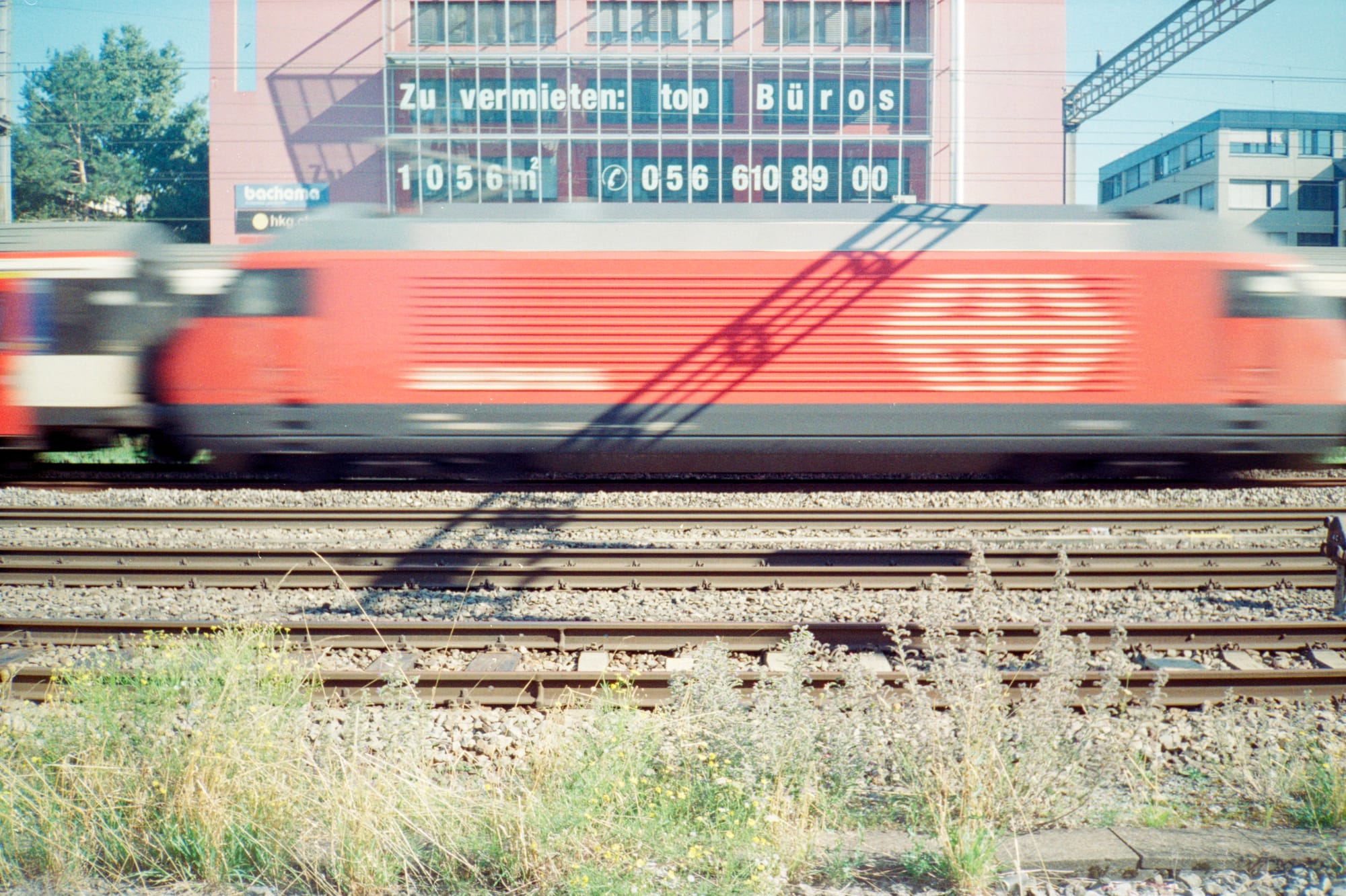 A red electric locomotive (SBB Re 460) thunders past, on a four track railway, hauling a passenger train. Overhead power stanchions cast a shadow across the loco. Behind, an industrious building with text in the windows: "Zu vermieten: Top Büros, 1056m²" plus a phone number.