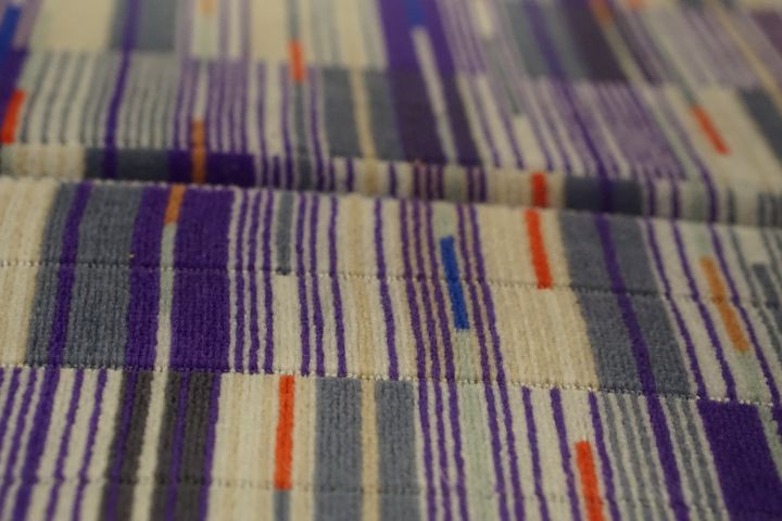 Close up of Elizabeth Line moquette on a train seat, with a white, grey, and royal purple pattern, with red and gold flashes.