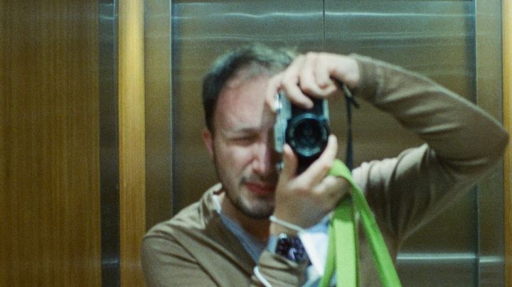 Jonathan in brown cardigan holding green reusable bag, taking a photo of himself in a lift mirror with an old SLR camera.