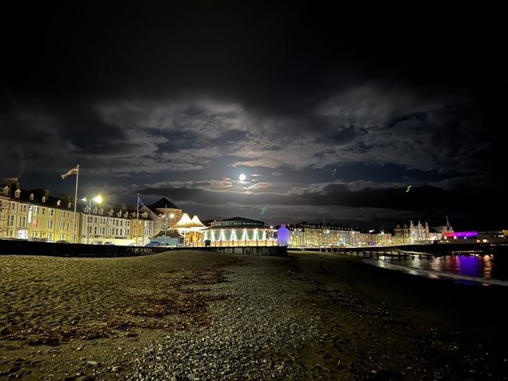 A shingle and sand beach at night with a promenade and a pier, lit by the moon between clouds.