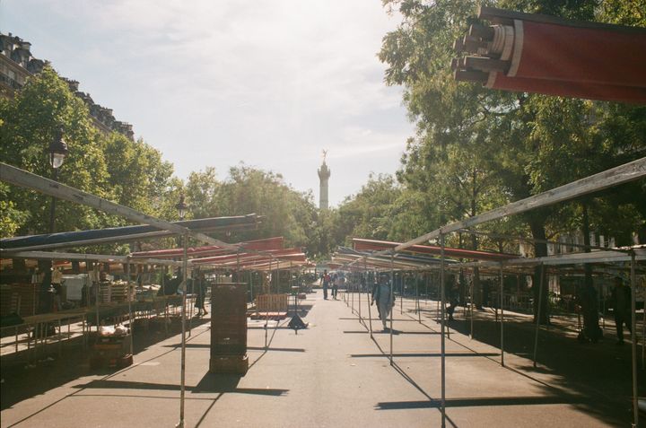 A marketplace being dismantled on a large area of asphalt, with rows of trees and a column in the distance in the sun