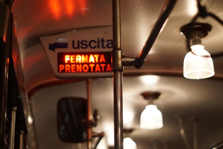Ceiling of a tram car with glass light fittings. A flashing red sign below the 'Uscita' sign reads "FERMATA PRENOTATA"