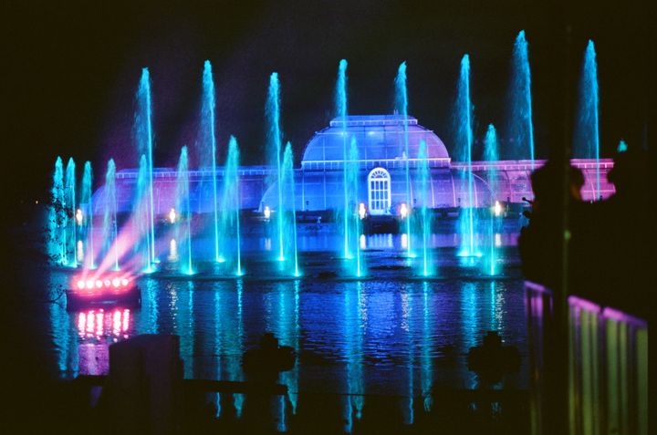 The Palm House at Kew Gardens with lit fountains in front, lit in bisexual lighting (blue, pink, purple.)
