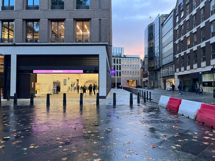 Outside Bond Street station (with illuminated purple sign) at dusk, with an orange sunset and wet yorkstone paving.