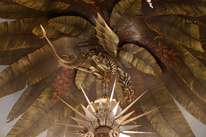 A ceiling with a palm leaf mural, with a giant metal dragon sculpture hanging above a sharded mirror chandelier. It’s A Lot.