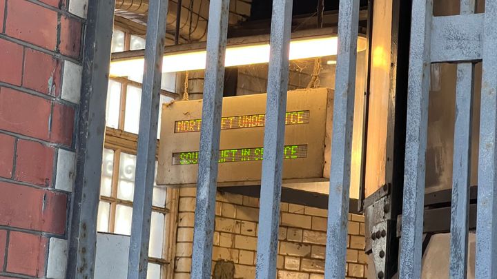 Into a building through a closed gate. A screen hanging from a ceiling says "NORTH LIFT UNDER MTCE, SOUTH LIFT IN SERVICE."