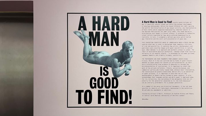 Wall with a text "A HARD MAN IS GOOD TO FIND!" with monochrome picture of a nude man (lying prone, from above) in the middle.