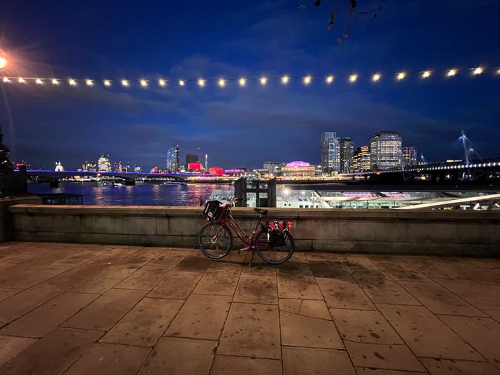 A red bicycle with a big crate on the front on a wide pavement in front of the River Thames at Embankment.