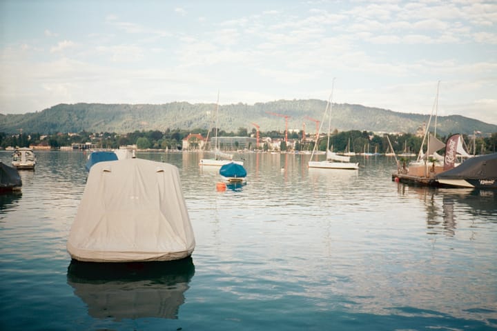 Small boats on a calm lake on a clear morning, little puffy clouds in the sky. Tree-covered hills rise in the distance.